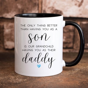 May include: White coffee mug with a black rim and handle. The mug has black text that reads "The only thing better than having you as a son is our grandchild having you as their daddy". A small blue heart is below the text.