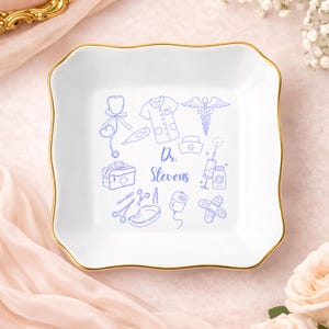 May include: White ceramic trinket dish with a gold rim and a scalloped edge. The dish features blue line art of medical symbols and the text "Dr. Stevens". Includes a stethoscope, scrubs, and a medical bag.