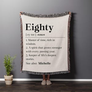 May include: A beige woven throw blanket with the word "Eighty" in large black letters. The blanket has a definition of the word "Eighty" printed below the word. The blanket has a fringed edge and is being held up by a person.