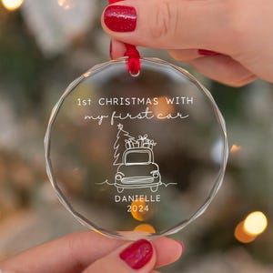 May include: Clear, faceted Christmas ornament with a red ribbon. The ornament features the text "1st CHRISTMAS WITH my first car" and a line drawing of a car carrying a Christmas tree and gifts. The name "DANIELLE" and the year "2024" are also included.