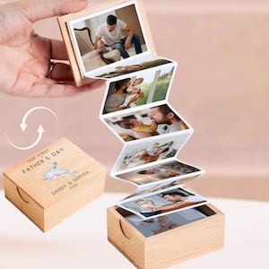 May include: A wooden photo box with a series of connected photos, held open to display the images. The box is engraved with "OUR FIRST FATHER'S DAY", a dolphin graphic, and "DADDY & SOPHIA 2026". The photos show a father and child.