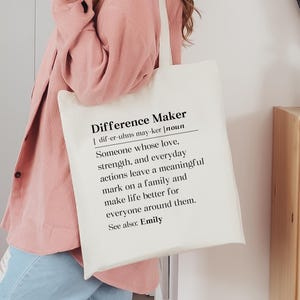 May include: A cream-colored tote bag with the text "Difference Maker" in black. The definition of "Difference Maker" is printed below the title. The bag is held by a person wearing a pink jacket.