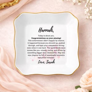 May include: A white ceramic trinket dish with a gold rim and a scalloped edge. The dish features the name "Hannah" and a congratulatory message. Pink floral accents are scattered across the text, and the message is signed "Love, Sarah."