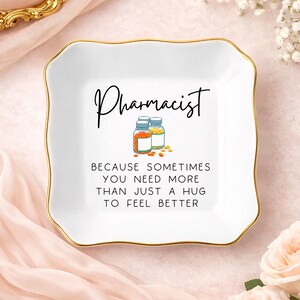 May include: A square white ceramic trinket dish with scalloped edges and a gold-painted rim. The center features the text Pharmacist, an illustration of two pill bottles with orange and yellow tablets, and the text BECAUSE SOMETIMES YOU NEED MORE THAN JUST A HUG TO FEEL BETTER.