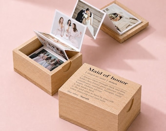 May include: A light-colored wooden box with a lid featuring the printed definition of Maid of honor. Inside the open box is an accordion-style photo album displaying multiple photographs of people. A second wooden box lid sits nearby.