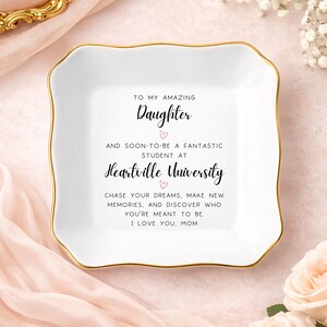 May include: A square white ceramic trinket dish with scalloped edges and a gold-painted rim. The center features black printed text that reads: TO MY AMAZING Daughter AND SOON-TO-BE A FANTASTIC STUDENT AT Heartville University CHASE YOUR DREAMS.