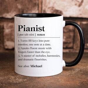 May include: White ceramic coffee mug with a black rim and handle. The mug features a black and white definition of the word "Pianist" with the pronunciation and a list of characteristics. The text at the bottom of the mug reads "See also: Michael."