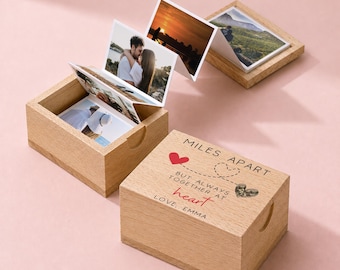 Könnte beinhalten: Eine helle Holzschachtel mit einem Deckel, der den eingravierten Text MILES APART BUT ALWAYS TOGETHER AT heart LOVE, EMMA trägt. Die Schachtel ist geöffnet und zeigt einen ziehharmonikaartigen Fotostreifen mit verschiedenen Bildern.