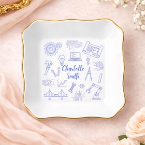 May include: White ceramic trinket dish with a gold rim and a square shape. The dish features blue line art of engineering tools and the name "Charlotte Smith". The design includes gears, a laptop, and a bridge.