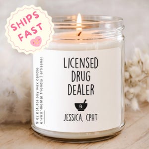 May include: A lit soy wax candle in a clear glass jar. The label reads "LICENSED DRUG DEALER" with a mortar and pestle symbol and "JESSICA, CPHT". The candle is 9 oz and is described as natural and artisanal.