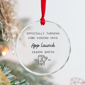 May include: A clear, round Christmas ornament with the text "OFFICIALLY TURNING VIBE CODING INTO App Launch" and "JASON SMITH". A red ribbon is attached to the top. The ornament has a smartphone graphic with a rocket ship.