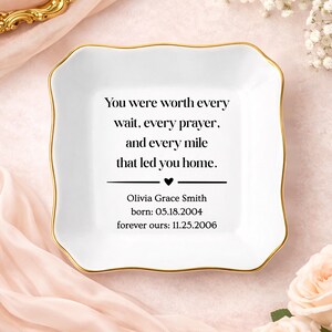 May include: A square white ceramic trinket dish with a gold-painted scalloped rim. The center features black text: You were worth every wait, every prayer, and every mile that led you home. Olivia Grace Smith, born: 05.18.2004, forever ours: 11.25.2006.