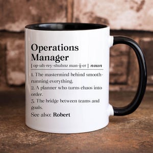 May include: White ceramic mug with a black rim and handle. The mug features a definition of "Operations Manager" with the phonetic spelling and three bullet points describing the role. The text at the bottom reads "See also: Robert".