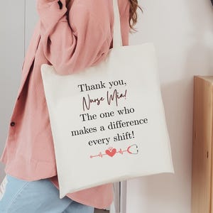 May include: Cream-colored tote bag with the text "Thank you, Nurse Mia! The one who makes a difference every shift!" in black and red. A red heart and stethoscope graphic is below the text.