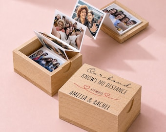 May include: A light-colored wooden box with a lid featuring the text Our bond KNOWS NO DISTANCE 479 MILES AMELIA & RACHEL. Inside the box is an accordion-style photo strip showing various group pictures of people, with another similar box nearby.
