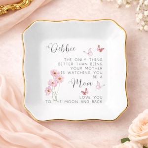 May include: White ceramic trinket dish with a gold rim. The dish features the name "Debbie" and a message about motherhood, along with pink floral and butterfly illustrations. The text reads "Love you to the moon and back."