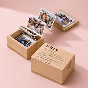 May include: Two light-colored wooden boxes with lids, one open displaying a pull-out accordion photo strip featuring various group portraits. The closed box lid is engraved with the text: Fifty | fif-tee | noun 1. Half a century of wisdom. 2. Shines with timeless charm and grace. 3. Inspires with a heart full of cherished moments. See also: Amelia.