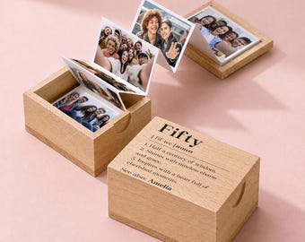 May include: Two light-colored wooden boxes with lids, one open displaying a pull-out accordion photo strip featuring various group portraits. The closed box lid is engraved with the text: Fifty | fif-tee | noun 1. Half a century of wisdom. 2. Shines with timeless charm and grace. 3. Inspires with a heart full of cherished moments. See also: Amelia.