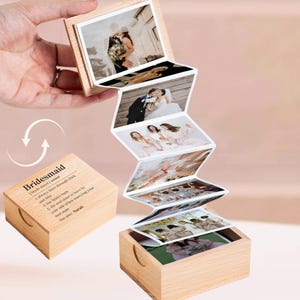 May include: A wooden box with a hinged lid that opens to reveal a mini photo album. The box is decorated with the word "Bridesmaid" and a definition of the word. The album contains several photos of a bride and her bridesmaids.