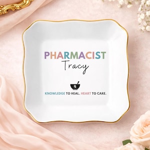 May include: A square white ceramic trinket dish with a gold-trimmed scalloped edge. The center features the text PHARMACIST in multicolored letters, the name Tracy in black script, a black mortar and pestle icon, and the text KNOWLEDGE TO HEAL. HEART TO CARE.