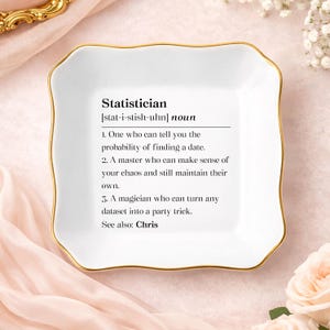 May include: A white ceramic trinket dish with a gold rim and a definition of "Statistician" printed in black. The definition includes three humorous points about the profession. The dish has a square shape with gently curved edges.