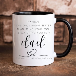 May include: White coffee mug with a black rim and handle. The mug has black text that reads "NATHAN, THE ONLY THING BETTER THAN BEING YOUR MOM IS WATCHING YOU BE A dad LOVE YOU TO THE MOON AND BACK".