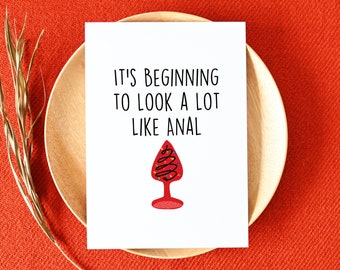 May include: A white greeting card with the text "IT'S BEGINNING TO LOOK A LOT LIKE ANAL" and a red Christmas tree-shaped design. The card is on a wooden plate, set against a red background.