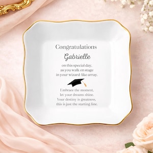 May include: A white ceramic trinket dish with a gold rim and a scalloped edge. The dish features the text "Congratulations Gabrielle" along with an inspirational message and a graduation cap illustration.