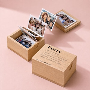 May include: Two light-colored wooden boxes with lids. One box is closed, displaying the printed text: Forty | fawr-tee | noun. 1. Masters the art of reinvention. 2. Bold life explorer, fearlessly embracing new horizons. 3. Dares to dream even bigger. See also: Michelle. The other box is open, revealing a pull-out accordion-style photo strip featuring various group portraits of people.