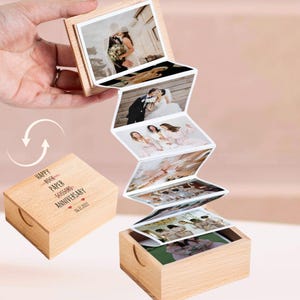 May include: A wooden photo box with a series of attached photographs, unfolding like an accordion. The box is engraved with the words "Happy Rock Paper Scissors Anniversary" and the date 06.12.2022. The photographs depict wedding scenes.