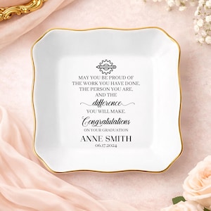 May include: A white, square trinket dish with a gold rim. The dish features a graduation message with the name "ANNE SMITH" and the date "06.17.2024". The text is in black, with a gear and pencil graphic at the top.