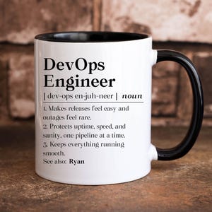 May include: A white coffee mug with a black handle and rim. The mug features the text "DevOps Engineer" with a definition and numbered points about the role. The mug is on a brown surface.