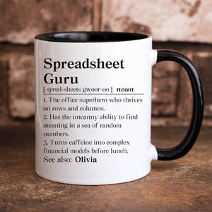 May include: White ceramic mug with a black interior and handle. The mug features the text "Spreadsheet Guru" with a definition, including the phrase "office superhero" and "Olivia".