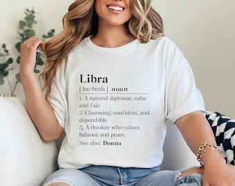 May include: White t-shirt with the definition of "Libra" printed in black. The definition includes the pronunciation, the word "noun", and three descriptive characteristics. The text also includes "See also: Donna".