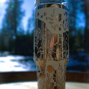 May include: A white and gold floral tumbler with a clear lid. The tumbler features a detailed design of flowers and foliage in gold and black. The background is blurred, showing a natural outdoor scene.
