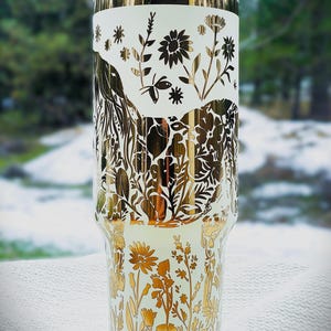 May include: A white and gold floral tumbler. The tumbler features a gold floral design with various flowers and leaves. The top and bottom of the tumbler have a gold floral pattern, while the middle section has a white background with gold floral accents.