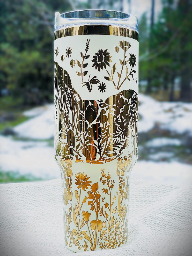 May include: A white and gold floral tumbler. The tumbler features a gold floral design with various flowers and leaves. The top and bottom of the tumbler have a gold floral pattern, while the middle section has a white background with gold floral accents.