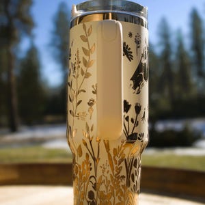 May include: Cream-colored travel mug with a clear lid and a white handle. The mug is adorned with a gold and black floral design. The handle is white. The mug is designed to keep beverages hot or cold.