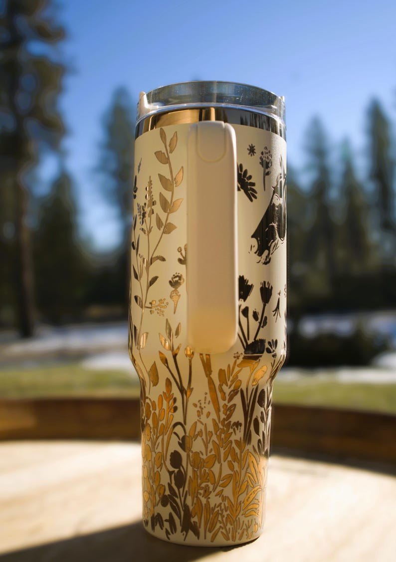 May include: Cream-colored travel mug with a clear lid and a white handle. The mug is adorned with a gold and black floral design. The handle is white. The mug is designed to keep beverages hot or cold.