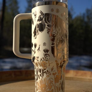 May include: Cream-colored insulated tumbler with a handle, featuring a gold and black horse and floral design. The tumbler has a clear lid and a cream-colored handle. The design includes a horse silhouette and various floral elements.
