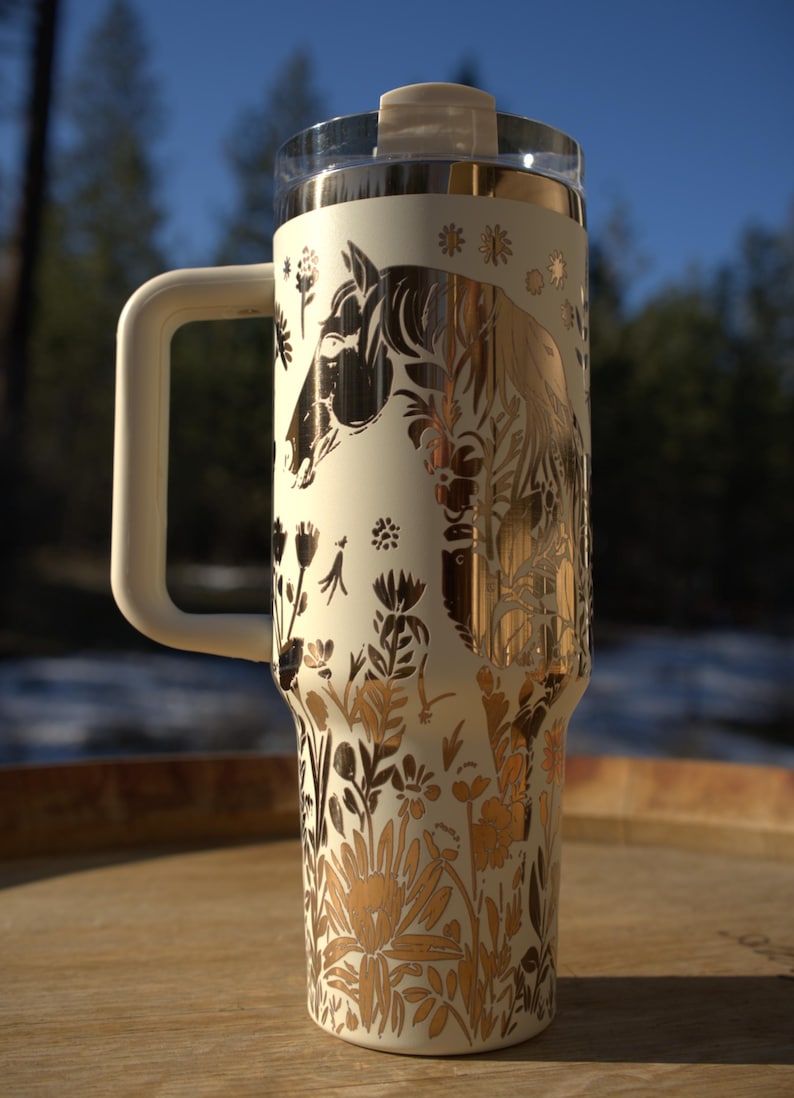 May include: Cream-colored insulated tumbler with a handle, featuring a gold and black horse and floral design. The tumbler has a clear lid and a cream-colored handle. The design includes a horse silhouette and various floral elements.