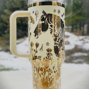 May include: Cream-colored insulated tumbler with a gold horse head and floral design. The tumbler has a cream-colored handle, lid, and accents. The design features a horse head silhouette and various flowers. The tumbler is designed to keep beverages cold or hot.