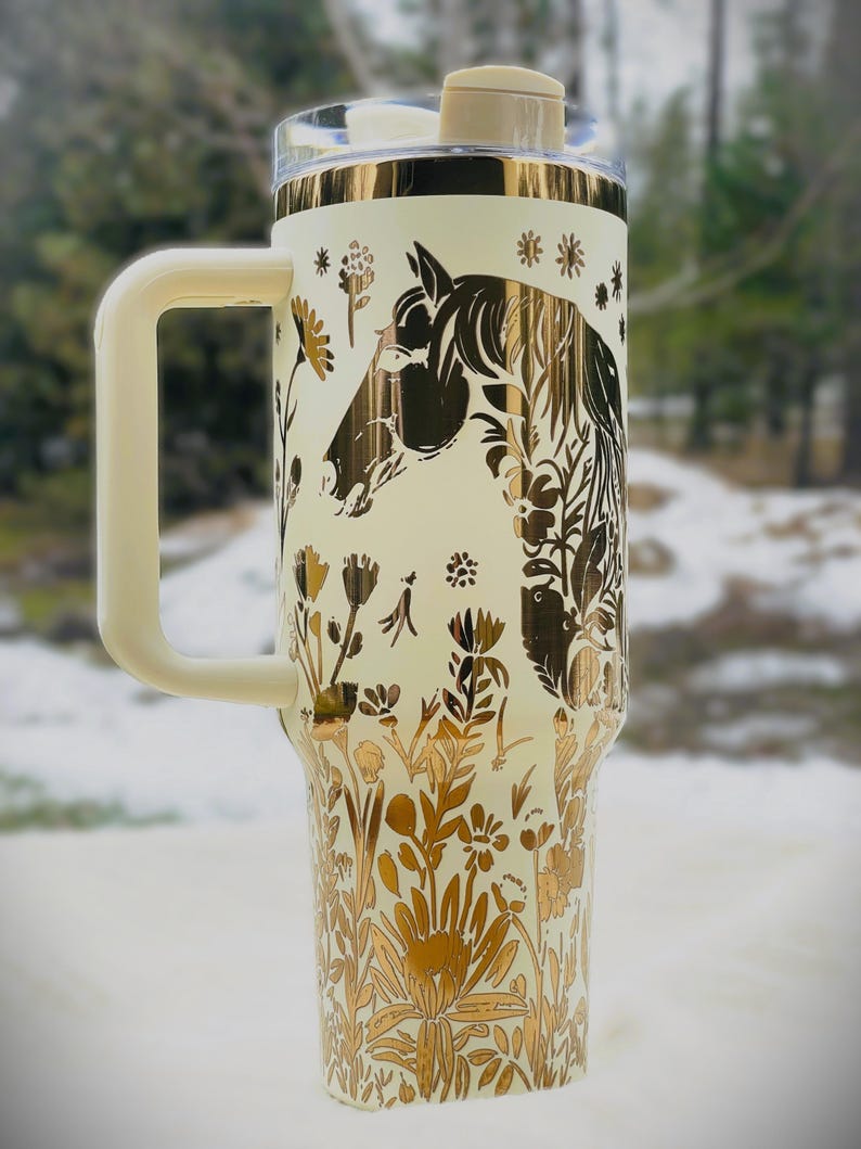 May include: Cream-colored insulated tumbler with a gold horse head and floral design. The tumbler has a cream-colored handle, lid, and accents. The design features a horse head silhouette and various flowers. The tumbler is designed to keep beverages cold or hot.