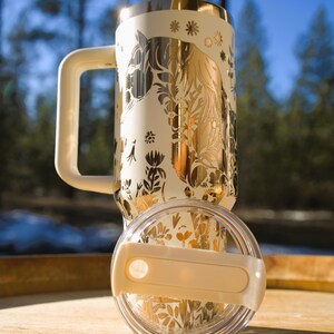 May include: Cream-colored travel mug with a gold horse and floral design. The mug has a matching handle and lid. The lid is clear with a cream-colored handle. The mug is sitting on a wooden surface.