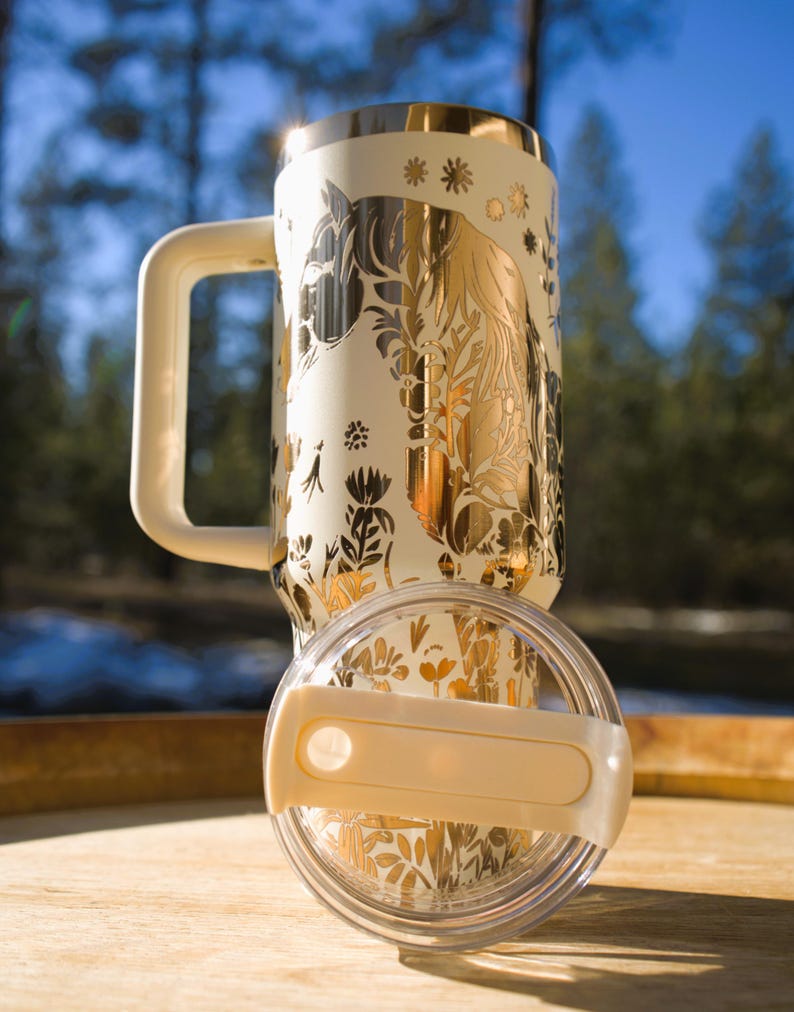 May include: Cream-colored travel mug with a gold horse and floral design. The mug has a matching handle and lid. The lid is clear with a cream-colored handle. The mug is sitting on a wooden surface.