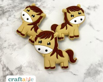 May include: Three brown and tan horse-shaped silicone charms with white eyes and brown manes. The charms are on a white marble surface.