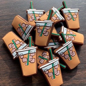 May include: A pile of brown and white iced coffee cups with green straws. The cups have a cartoon-like design with ice cubes and a brown liquid inside.