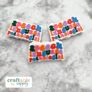 May include: Three white, cloud-shaped charms with the words "Speech Language Pathologist" in colorful, bubble letters. The charms are in shades of green, orange, purple, and blue. The background is a light gray, textured surface.