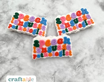 May include: Three white, cloud-shaped charms with the words "Speech Language Pathologist" in colorful, bubble letters. The charms are in shades of green, orange, purple, and blue. The background is a light gray, textured surface.