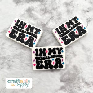 May include: Three white rectangular charms with the text "IN MY CHEERLEADER ERA" in black, with colorful accents. The charms have a retro, groovy aesthetic, perfect for crafting or personalizing accessories.